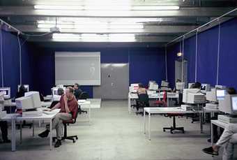 Photograph of a large room with blue walls and concrete floors, lit by bright fluorescent strip-lights on the ceiling. There are a number of white office desks with a desktop computer on each. Some people sit on red swivel chairs at the computers.