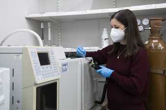 A person wearing a white face mask and blue protective gloves operates a piece of machinery. They are in a lab, surrounded by large machines and cylinders.