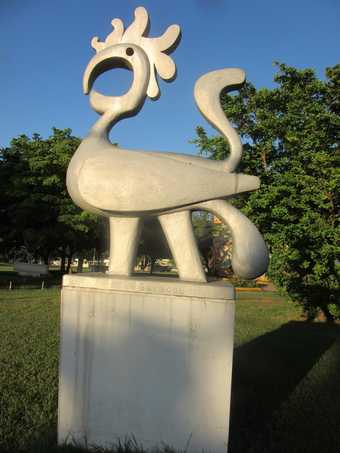 Photograph of an aluminium sculpture of a stylised bird on a square pedestal (on which the title Savacou is inscribed), installed in a park with grass, trees and blue sky in the background