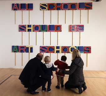 A family of two parents and two children are looking at an artwork by Bob and Roberta Smith which says 'All Schools Should Be Art Schools'