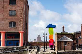 the side of Tate Liverpool, a red brick industrial building held up by large red pillars. To the right is a large sculpture made up of brightly coloured rocks stacked vertically. The sky behind is blue with white fluffy clouds.
